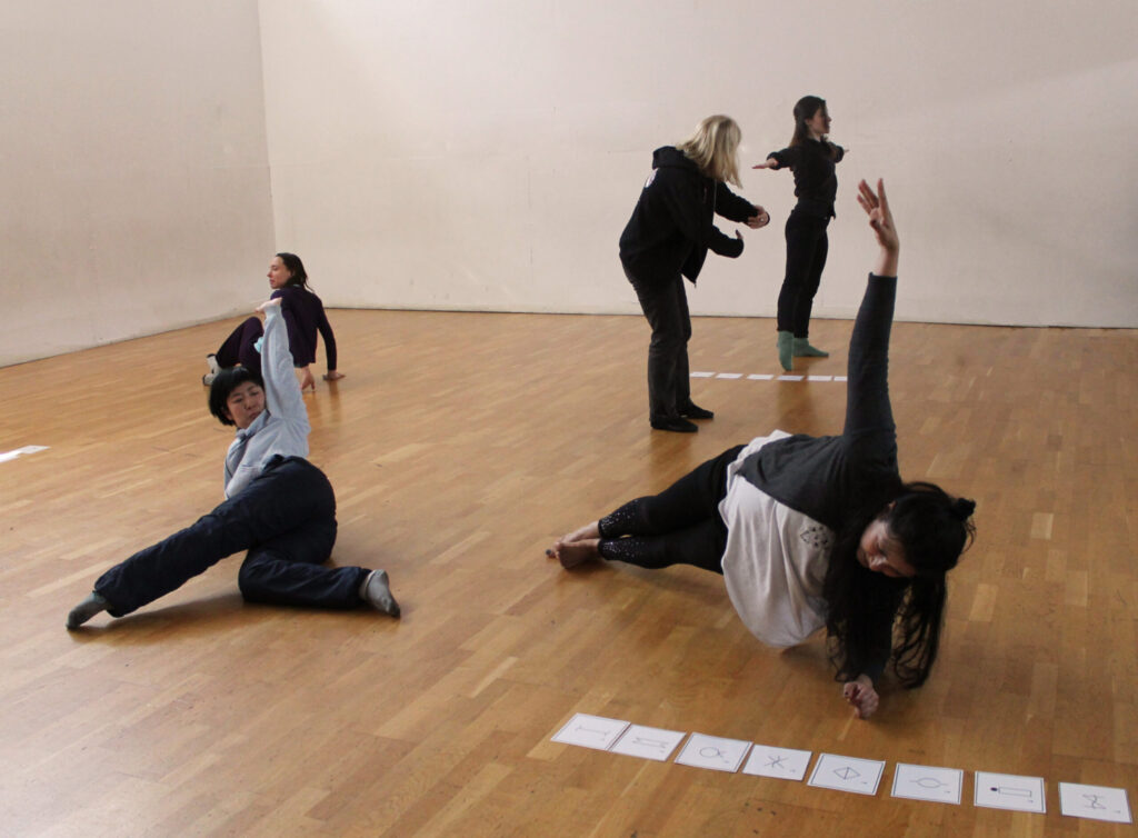 Group of adults in a dance studio holding different poses and looking at symbols flashcards laid out on the floor.