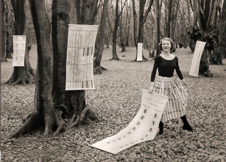 Black and white picture of Dr Ann Hutchinson Guest in a forest surrounded by notation scores hung on tree trunks