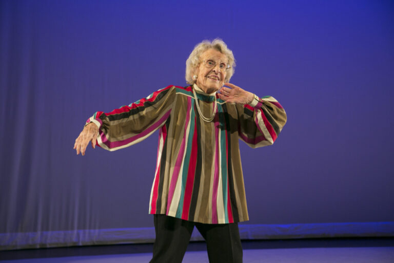 Dr Ann Hutchinson Guest in a brightly coloured shirt, smiling and striking a pose on a lit stage